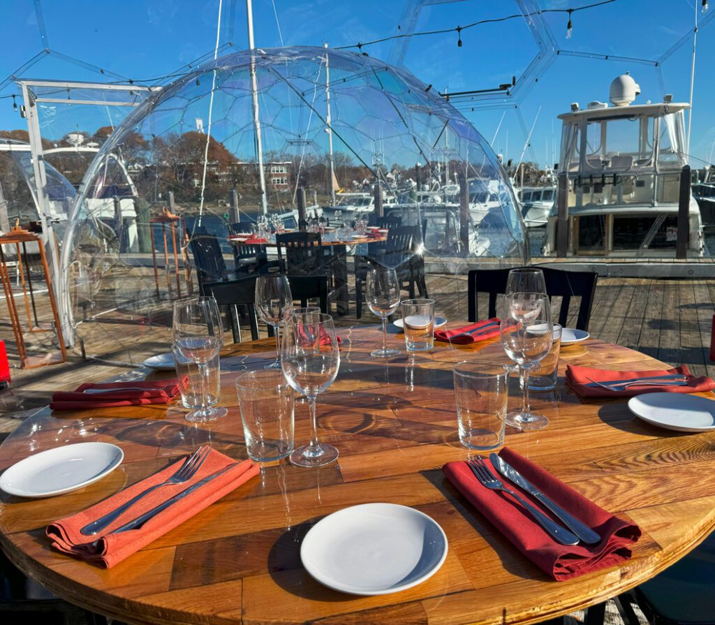 Clear dome dining table chairs glowing light perimeter at daytime overlooking a marina and boats