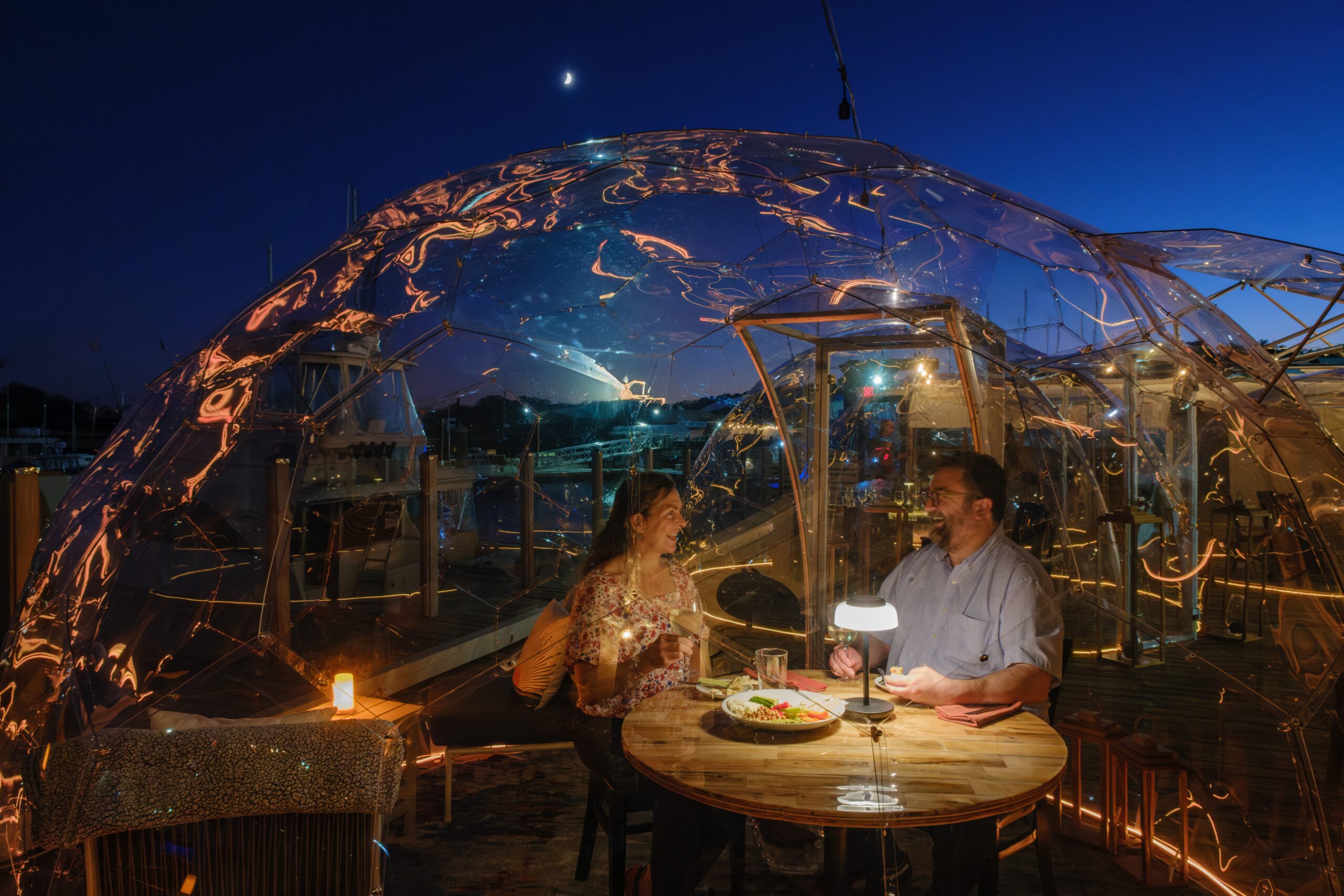 woman and man dining in a clear igloo with boats in the background marina
