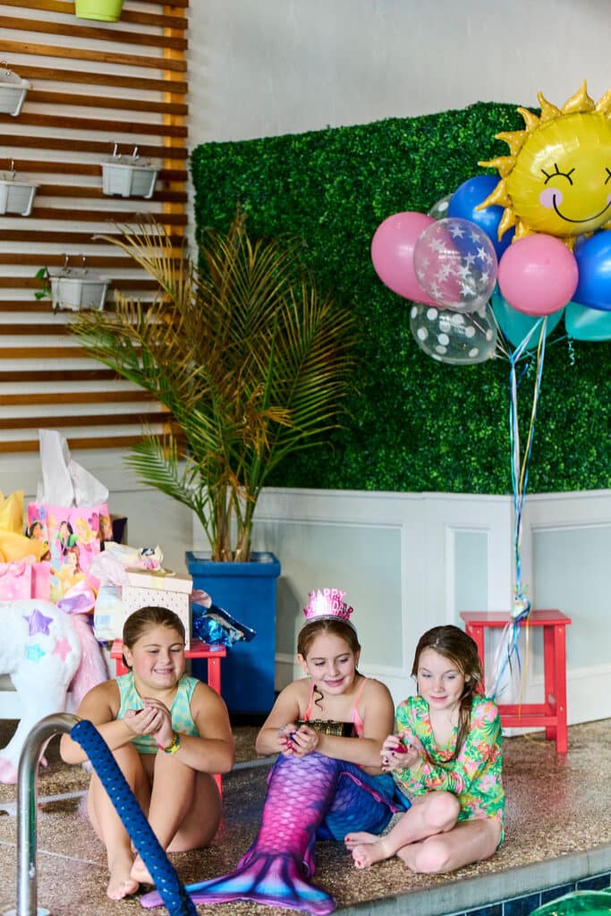 Three girls sitting next to a pool with ballons, one has a mermaid tail