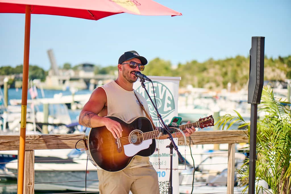 Guy playing guitar with baseball cap sining at microphone with marina boats in background