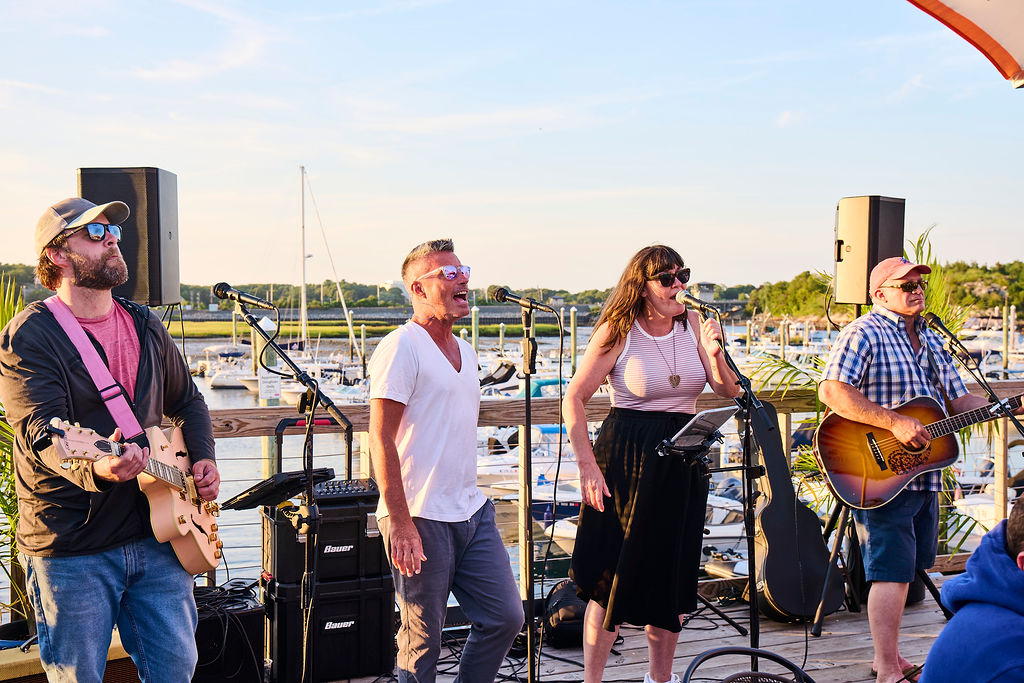 men and women outside on deck wiht marina in background singing and playing music band