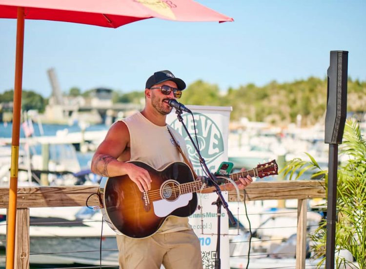 man playing guitar outside boats in the background sining
