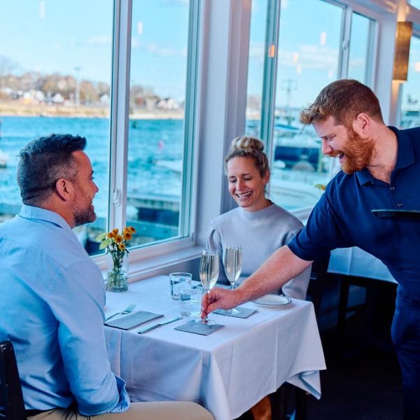 male and female getting served champagne at a table with marina water in background at a restaurant