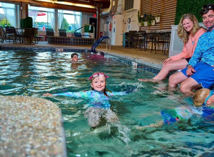 girl in swimshirt goggles on head in pool with parents watching