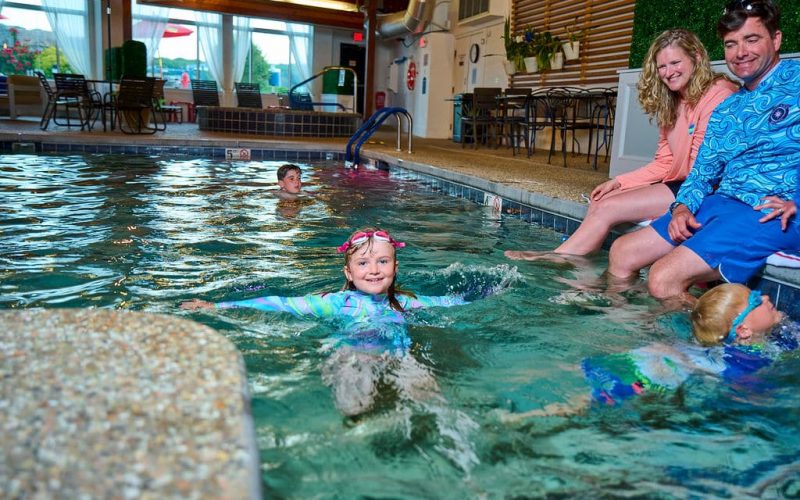 girl in swimshirt goggles on head in pool with parents watching