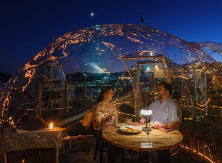 woman and man dining in a clear igloo with boats in the background marina