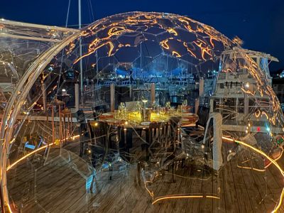 Clear dome dining table chairs glowing light perimeter at night overlooking a marina and boats