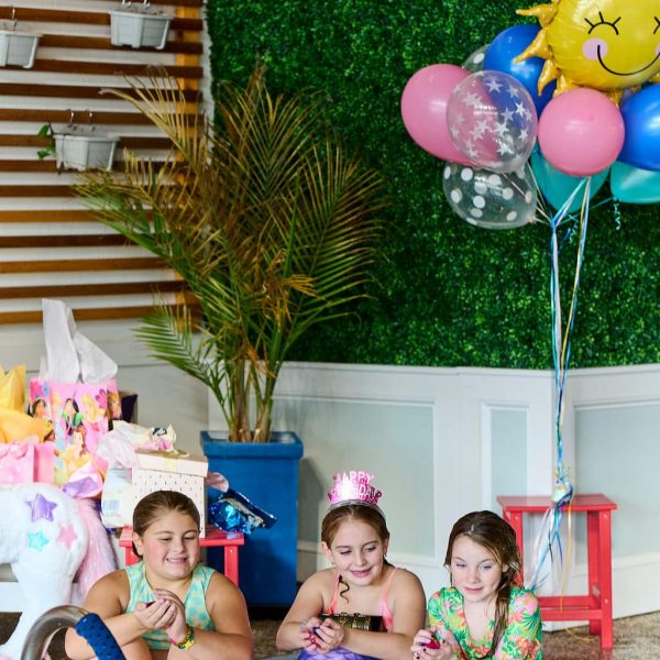Three girls on the side of an indoor pool, one with mermaid tail on