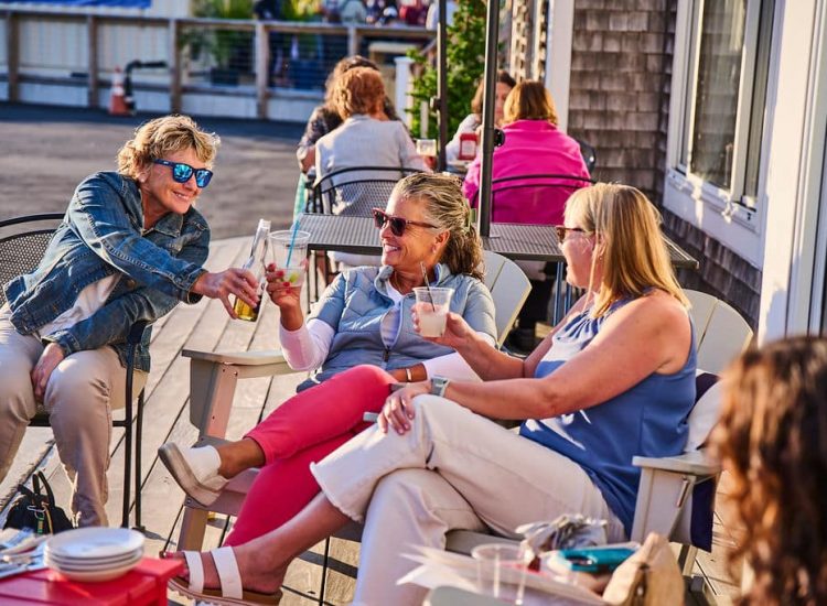 Ladies in teh sun sitting with sunglasses cheers with drinks