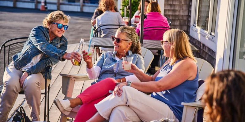 Ladies in teh sun sitting with sunglasses cheers with drinks